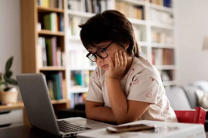 Cute boy with hand on chin looking at laptop while sitting in living room