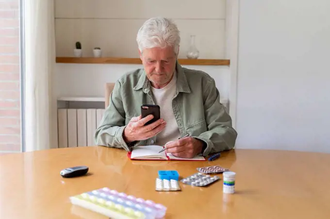 Senior man using smart phone while sitting with diary and medicines on table at home