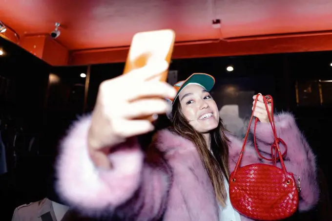 Happy woman taking selfie through smart phone while wearing fur jacket and holding leather purse in store