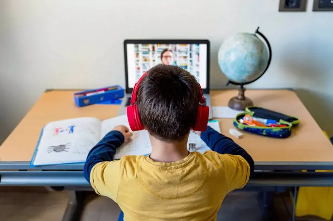 Rear view of boy listening to teacher through video call at home during pandemic outbreak