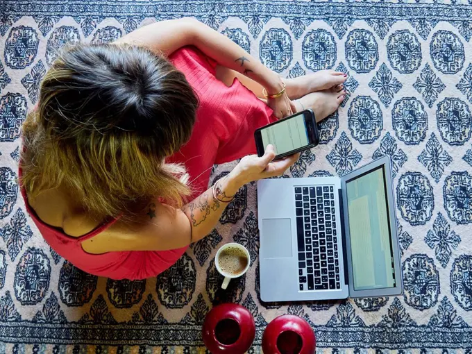 Woman sitting on the floor with cup of coffee and laptop using smartphone