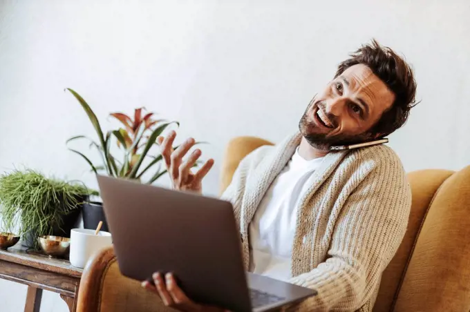 Portrait of man on the phone sitting on armchair with laptop pulling funny faces