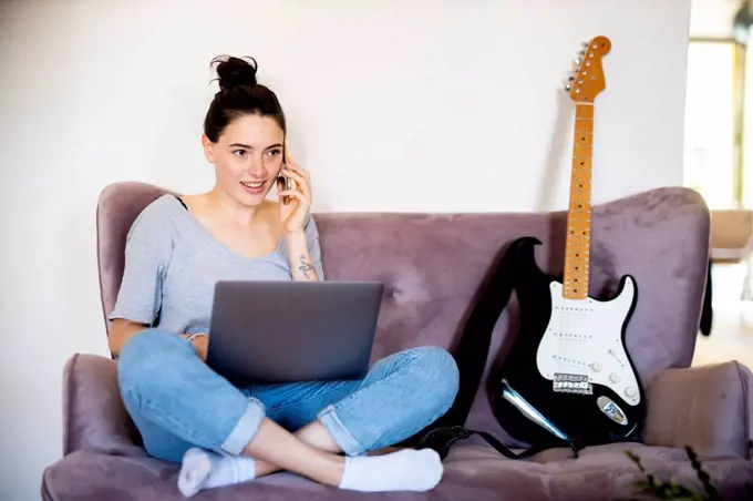 Portrait of young woman on the phone sitting with laptop on couch at home