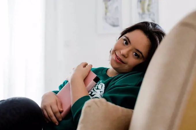 Smiling young woman with down syndrome holding book while resting on armchair at home