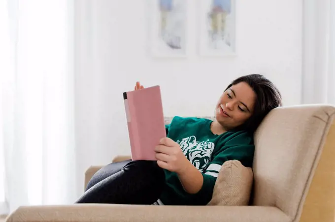 Happy young woman with down syndrome reading book while resting on armchair at home