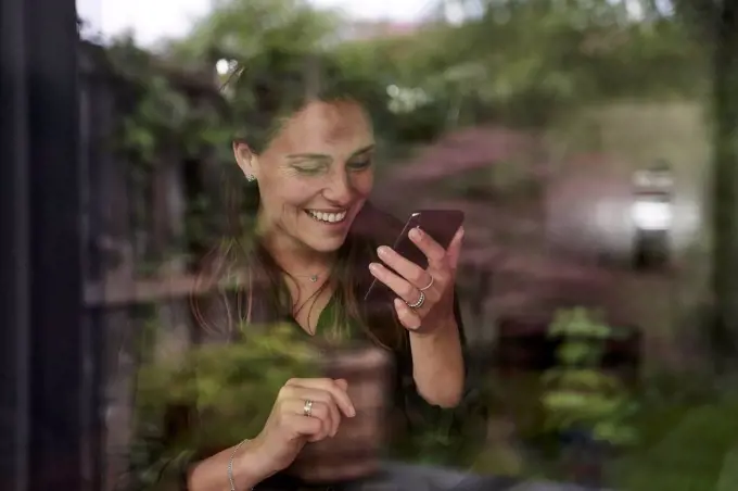 Smiling mid adult woman using mobile phone at home seen through window