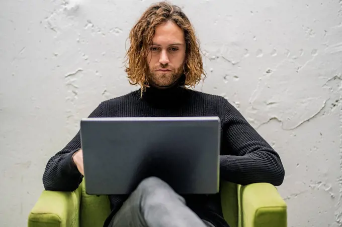 Bearded young man using laptop while sitting on armchair against wall at home