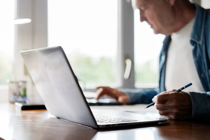 Senior man using calculator and laptop while writing at home