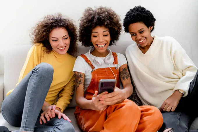 Group picture of three laughing friends sitting on couch taking selfie with smartphone