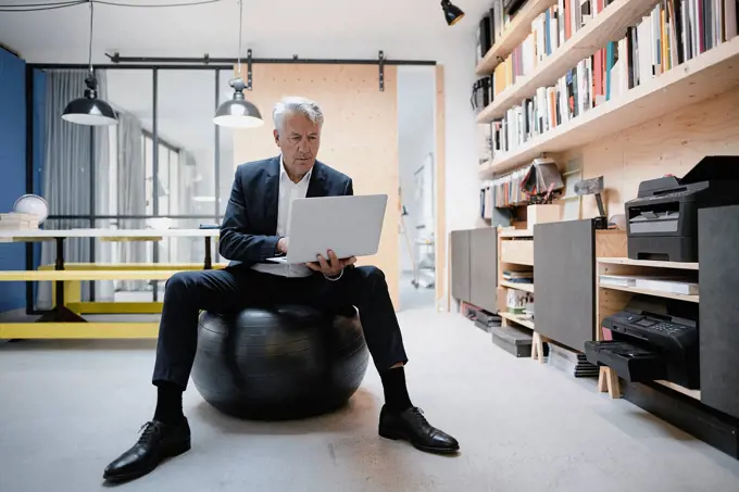 Senior businessman sitting on fitness ball, using laptop