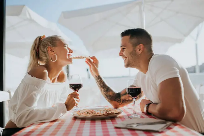 Happy young man feeding pizza to girlfriend while sitting in restaurant