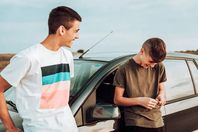 Male friends laughing while standing by car against sky at sunset