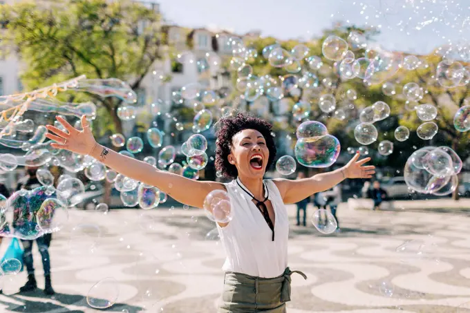 Carefree mid adult woman playing with bubbles while standing in city