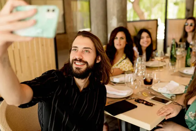 Smiling young man taking selfie with female friends sitting at table in restaurant