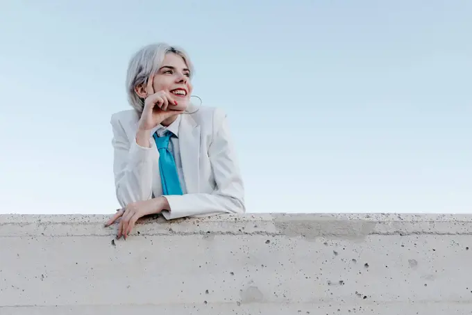 Smiling young woman wearing white suit standing by retaining wall against clear sky