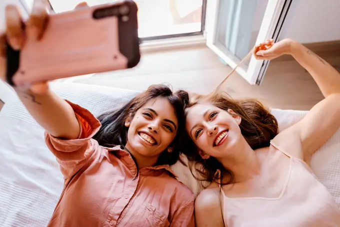 Friends taking selfie with mobile phone while lying on bed at home