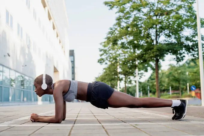 Female athlete listening music while practicing planks on footpath in city