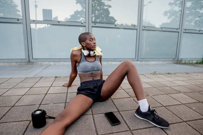 Tired female athlete with shaved head sitting on footpath in city