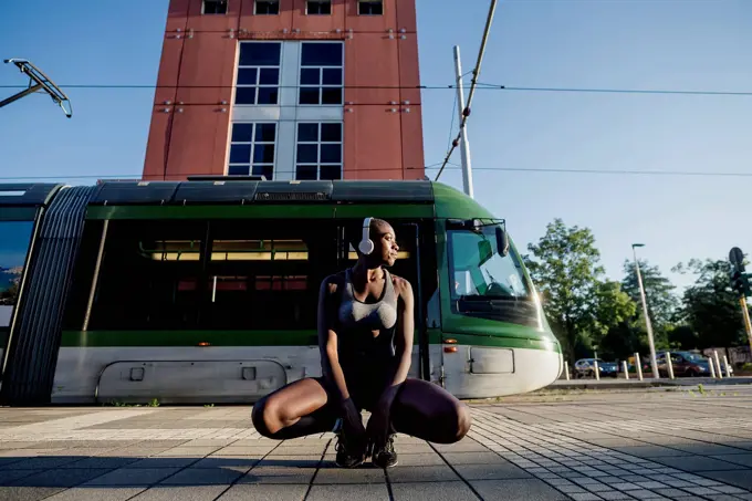 Female athlete listening music through headphones while crouching on street in city