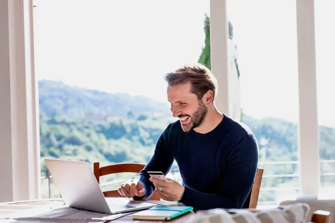 Smiling handsome man holding credit card while using laptop at dining table against window