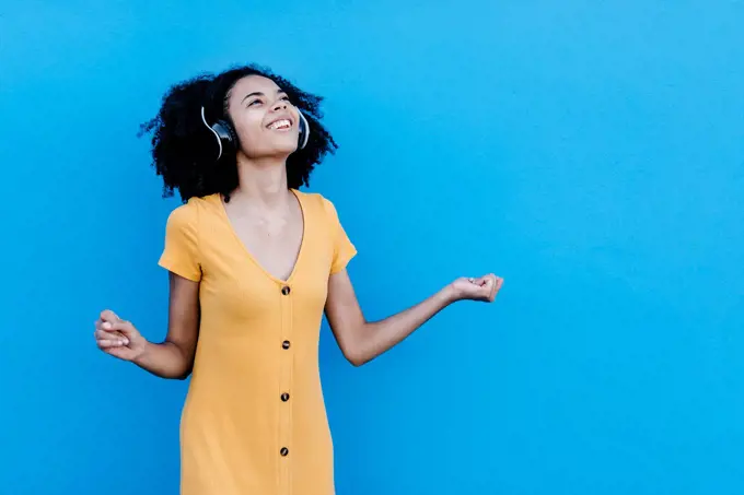 Happy young afro woman enjoying while listening music through headphones against blue wall