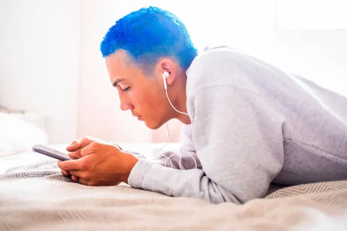 Young man with blue hair using smartphone and listening to music lying on bed