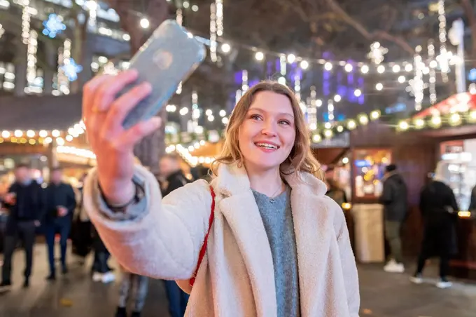 Young woman taking selfie while standing in illuminated Christmas market at night