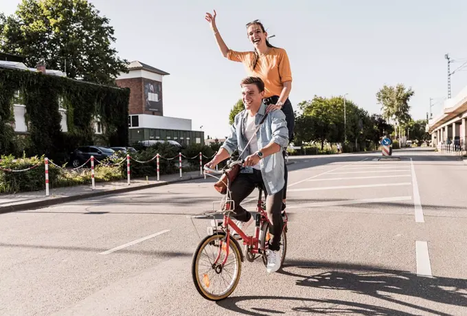 Cheerful young woman standing behind boyfriend riding bicycle on street