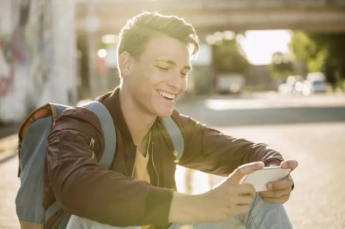 Happy young man using smart phone while sitting on street during sunny day