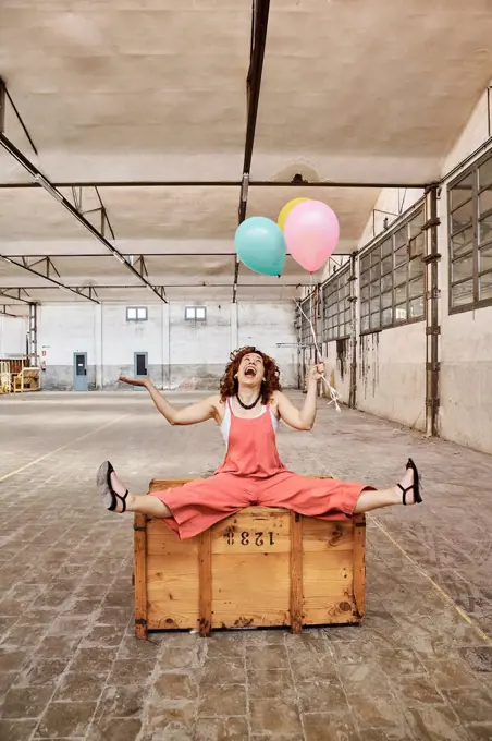 Cheerful woman playing with colorful helium balloons while sitting on wooden box