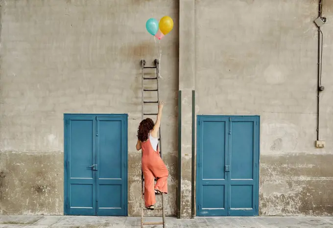 Woman climbing ladder leaning on wall while reaching for colorful helium balloons