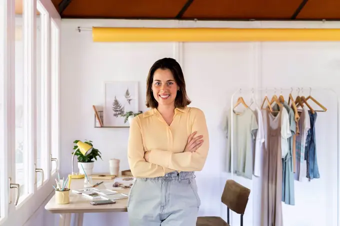 Smiling young female fashion designer with arms crossed leaning on table at clothing store