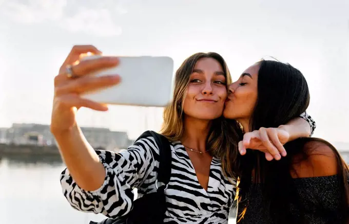 Young woman kissing friend while taking selfie against clear sky