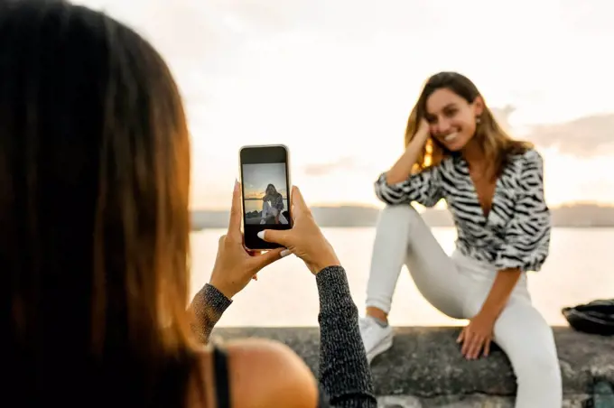 Woman taking photo of friend sitting on retaining wall at promenade