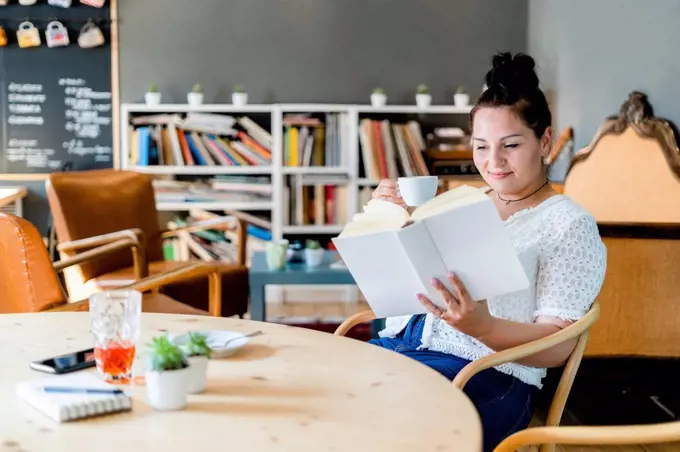 Young woman holding coffee cup reading book while sitting at table in cafe