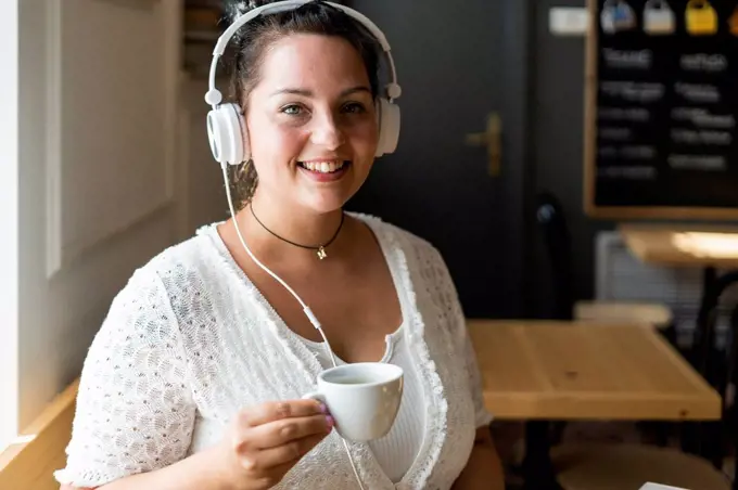 Close-up of smiling young woman holding coffee cup listening music through headphones in cafe