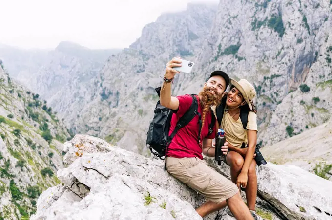 Smiling couple taking selfie while sitting on rock at Ruta Del Cares, Asturias, Spain