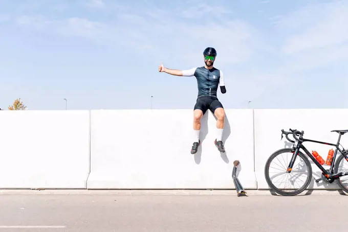 Male amputee athlete sitting on retaining wall against sky during sunny day