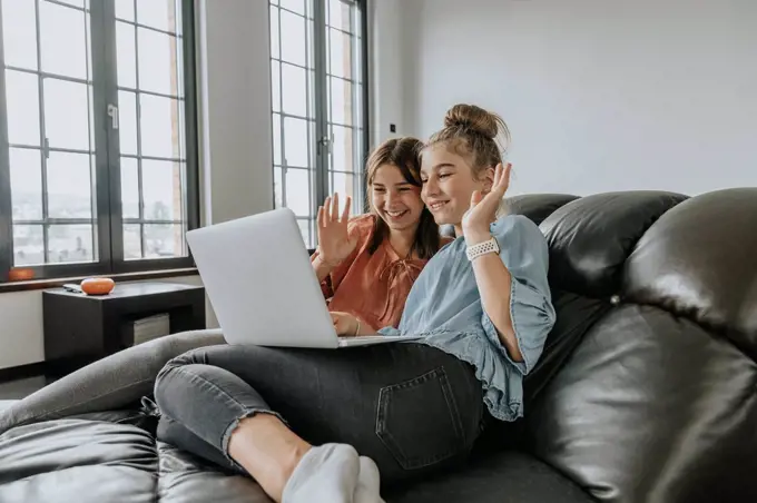 Friends waving while video conferencing over laptop on sofa at home