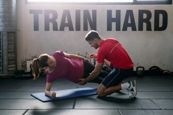 Fitness instructor assisting female athlete in practicing plank position on mat at gym