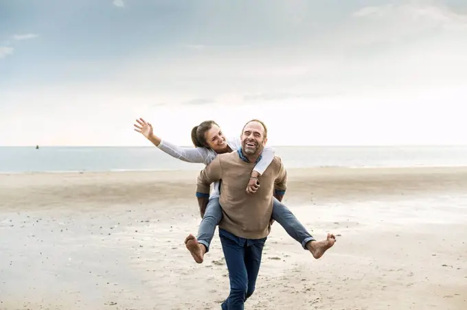 Happy mature man piggybacking woman while walking at beach against cloudy sky