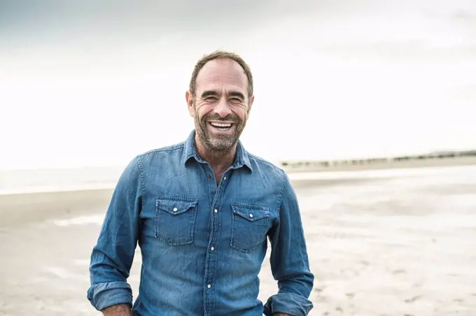 Cheerful man laughing while standing at beach against sky during sunset