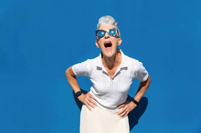 Woman with mouth open and hand on hip standing against blue wall during sunny day