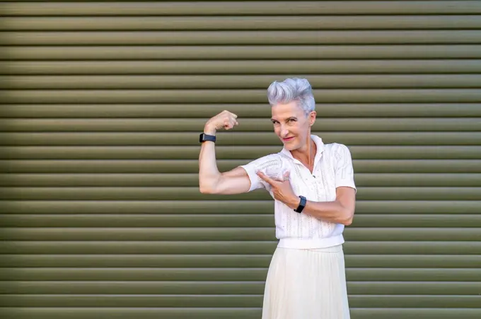 Active senior woman pointing at bicep while flexing muscles while standing against shutter