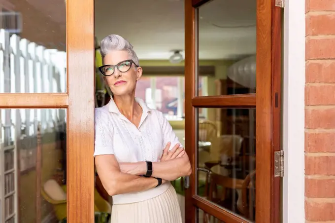 Contemplating senior woman with arms crossed standing on doorway at home