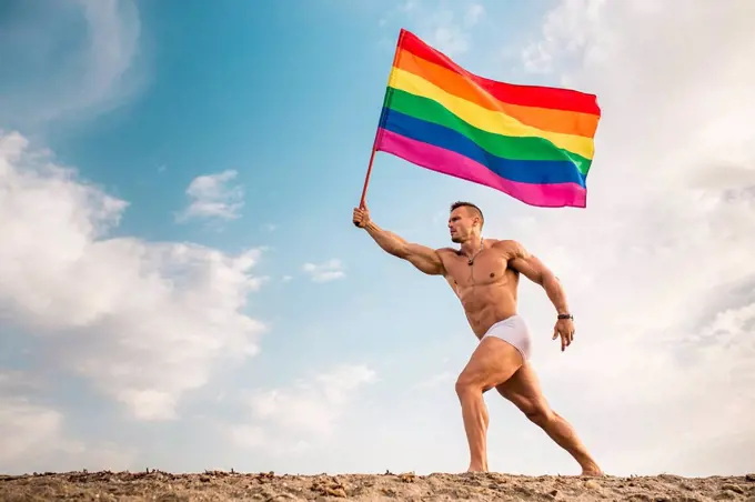 Shirtless young man holding rainbow flag walking at beach against sky