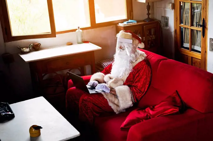 Man wearing Santa Claus costume using laptop while sitting on sofa at home