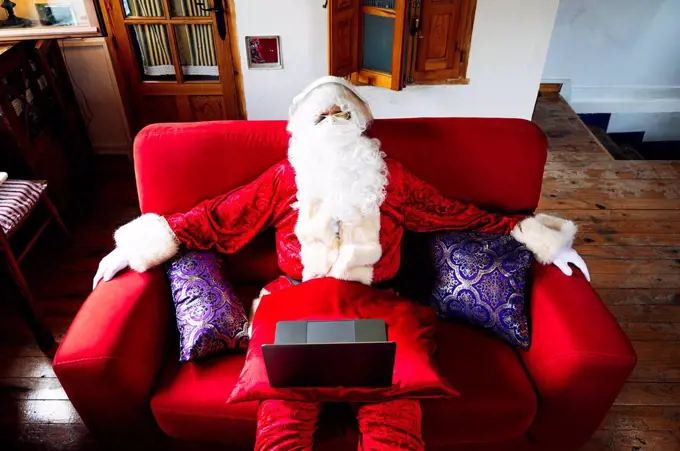 Man wearing Santa Claus costume with laptop relaxing on sofa at home