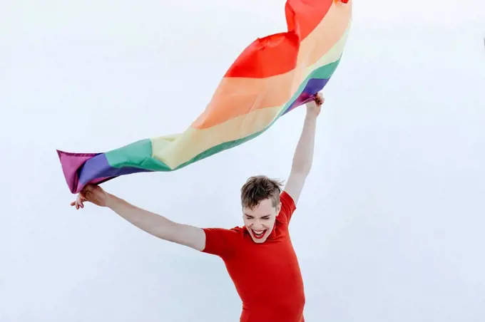 Non-binary person waving multi colored flag while standing against white wall