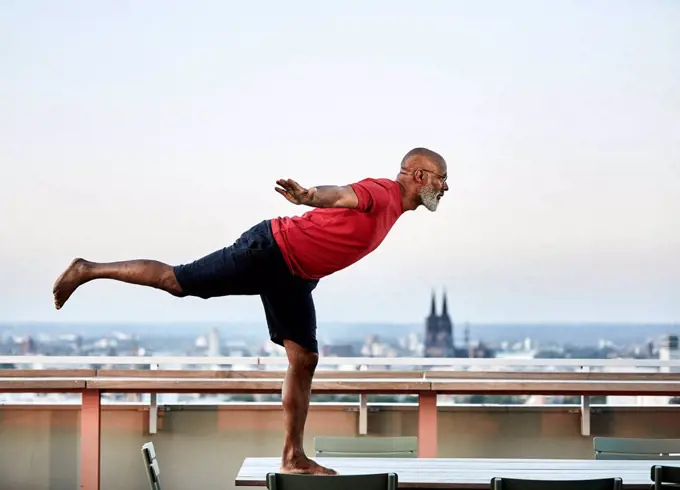 Bald man balancing on dining table while practicing yoga at building terrace during sunset